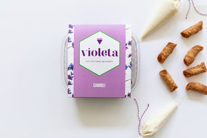 Lavender box of gluten free cannoli shells, surrounded by six cannoli shells and two piping bags of filling, tied with purple and white baker's string.
