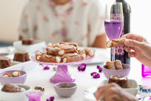 Load image into Gallery viewer, Two women sit at a table filled with gluten free sweets, fresh flowers, a bottle of wine and glasses. One woman holds a gluten free cannoli in her hand.