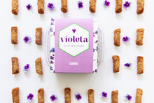 Load image into Gallery viewer, Lavender box of gluten free cannoli, surrounded by empty cannoli shells and violets in an alternating pattern.