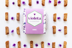 Lavender box of gluten free cannoli, surrounded by empty cannoli shells and violets in an alternating pattern.