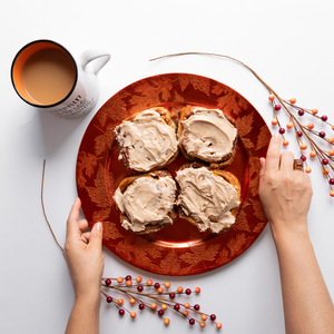 Overhead shot of woman placing orange plate of cinnamon rolls on a table. Mug of coffee next to the plate, and decorative branches are on the table.
